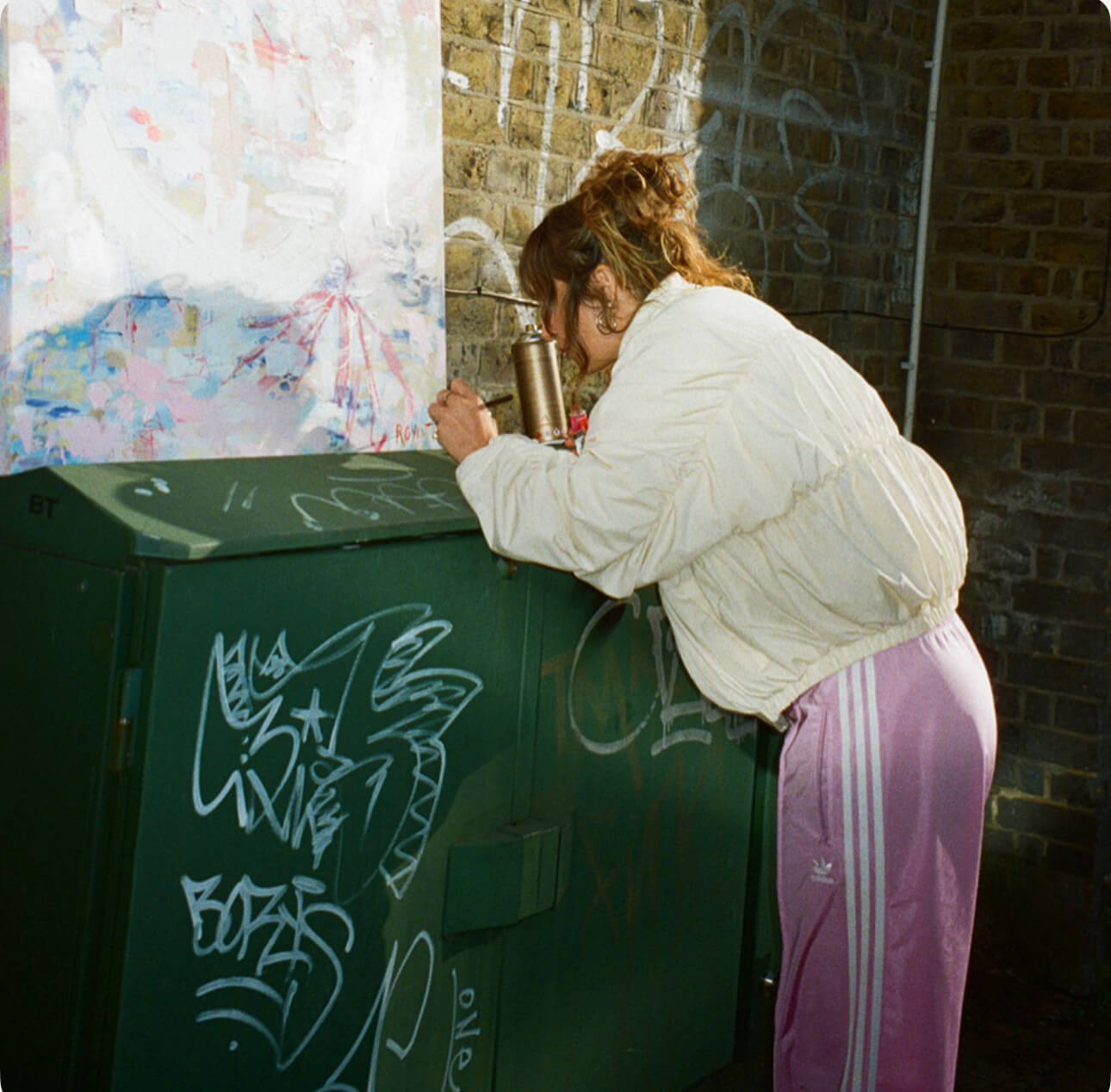 Sara painting on a green board with graffiti against a brick wall.