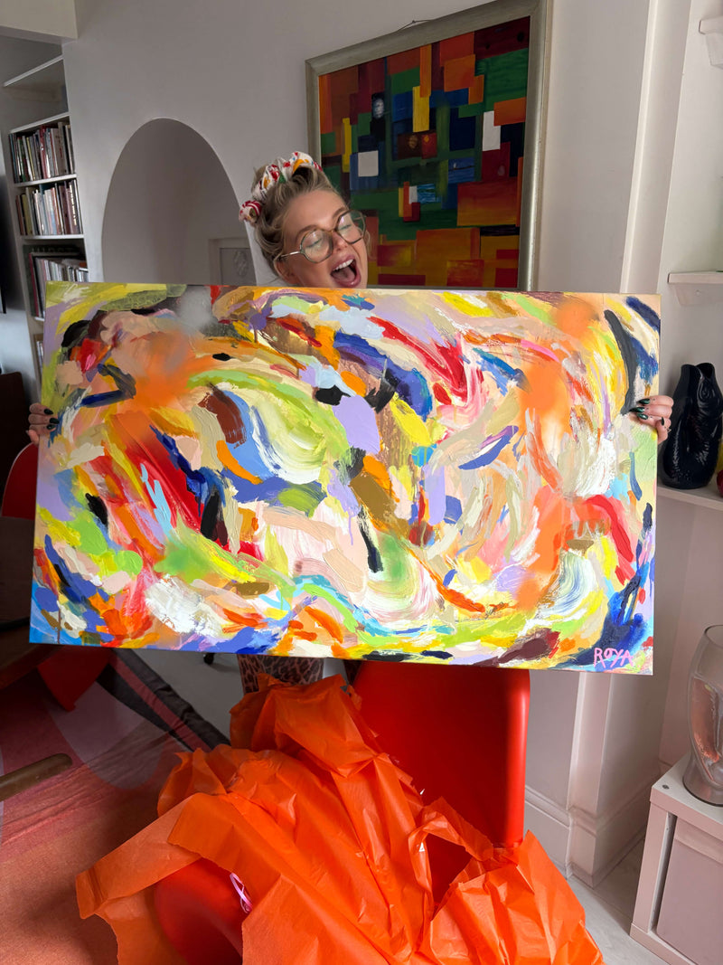 Person holding a colorful abstract painting in a room with a bookshelf and another artwork on the wall.
