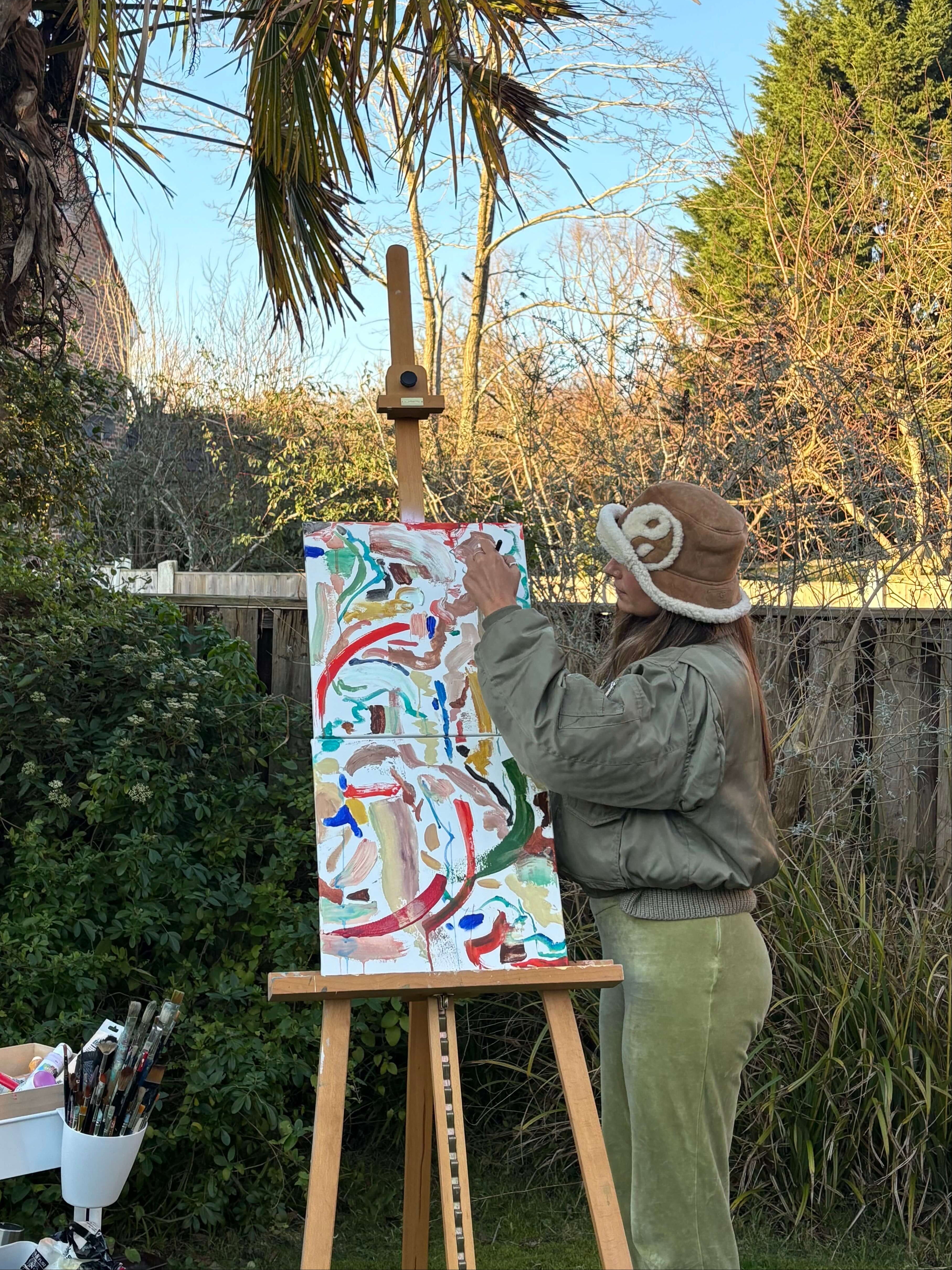 Sara painting on an easel in a garden setting with trees and a clear sky.